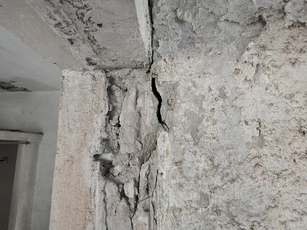 An open, near-vertical crack running down the junction between a projecting vertical element and the adjoining wall, with the surface finish broken away in a strip alongside it and heavy pitting across the adjacent wall, in a Puerto Rico building